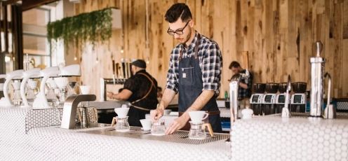 A barista prepares coffee with pour-over equipment in a cafe, wood paneling behind him.