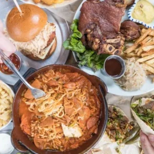 Overhead shot of various dishes: sandwich, pork knuckle, pasta, fries, tacos, and sauce bowls.