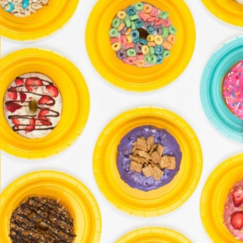 Donuts with various colorful toppings, arranged on yellow and blue paper plates, on a white surface.