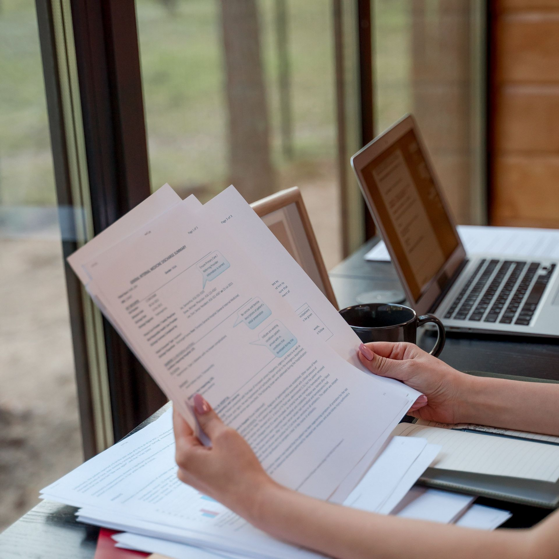 Hands reviewing printed documents beside a laptop on a desk by a window