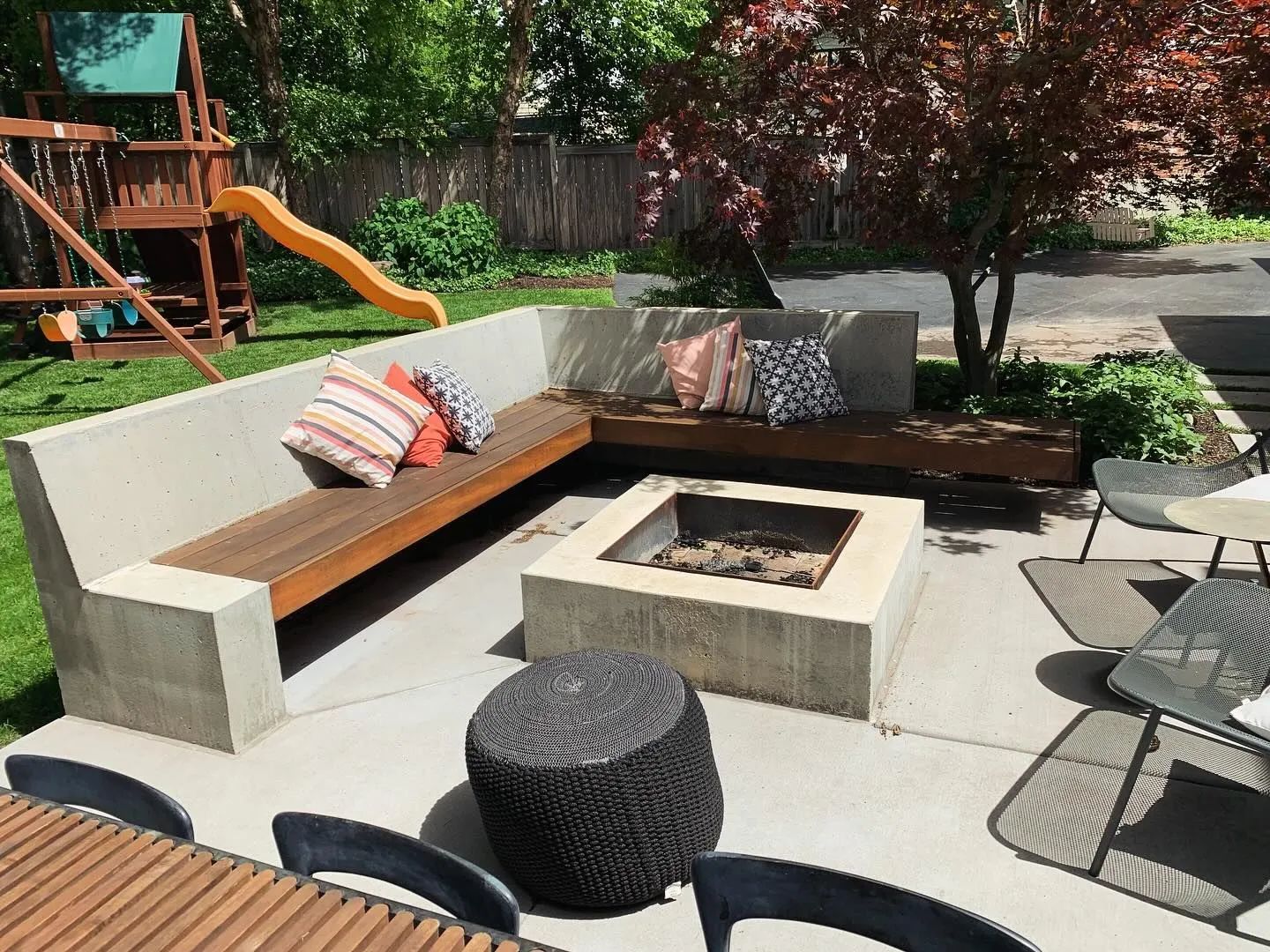 Outdoor patio with L-shaped bench seating, fire pit, and string chairs in a backyard garden