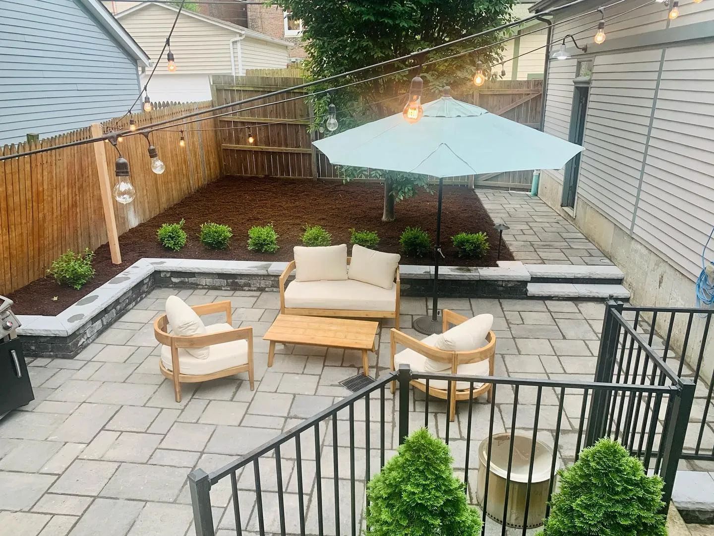 Backyard patio with wicker seating, a blue umbrella, planters, and string lights at dusk