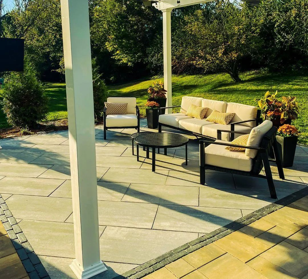 Patio with outdoor furniture under a white pergola, set on patterned gray and yellow stone.