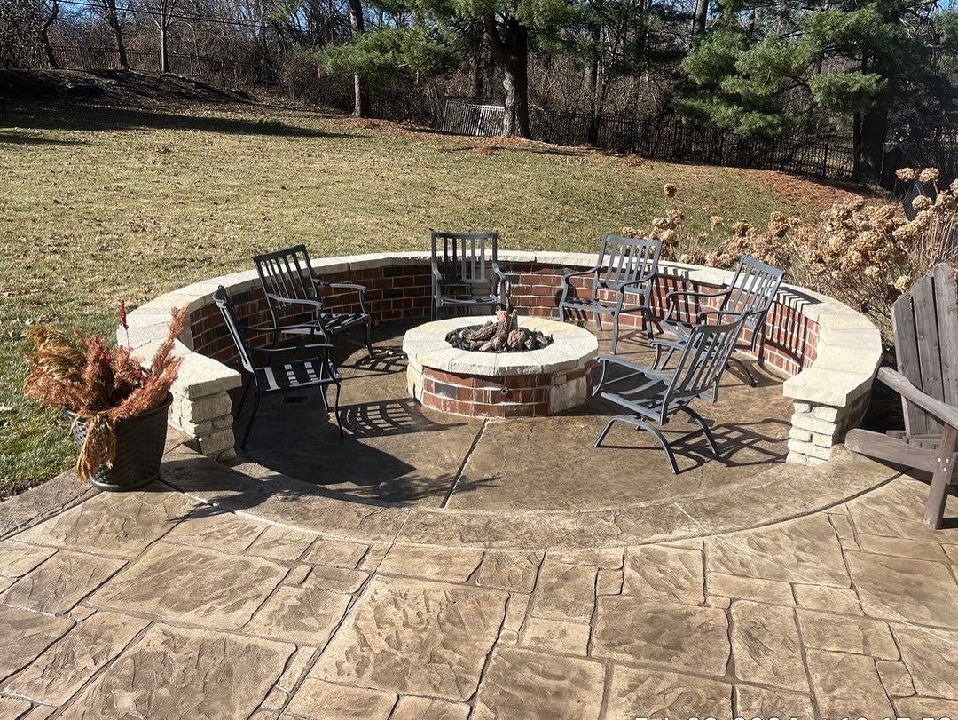 Circular brick fire pit surrounded by metal chairs on a stone patio in a backyard setting.