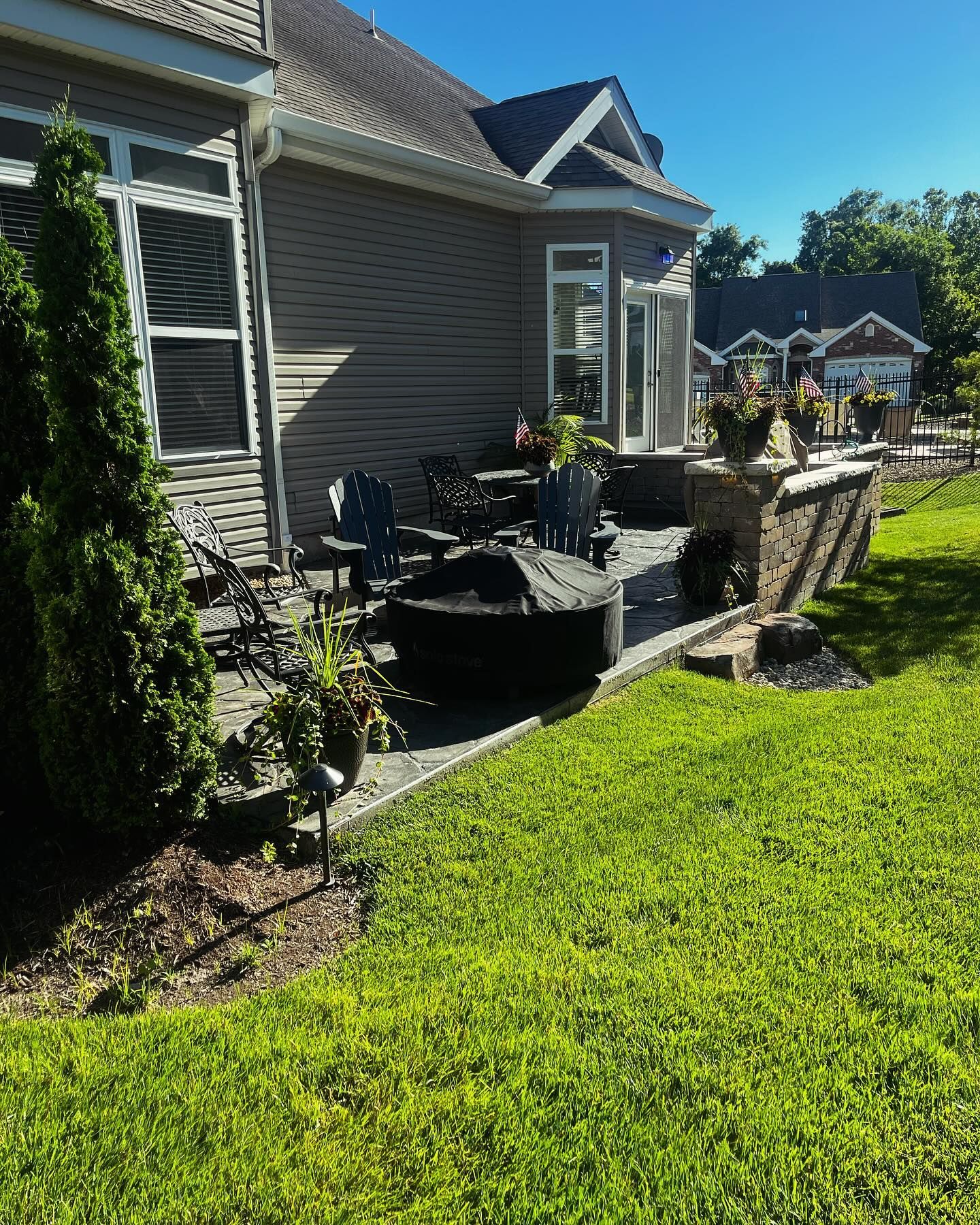 Backyard patio with a fire pit, seating, and potted plants, surrounded by a low retaining wall and lush green grass, 
