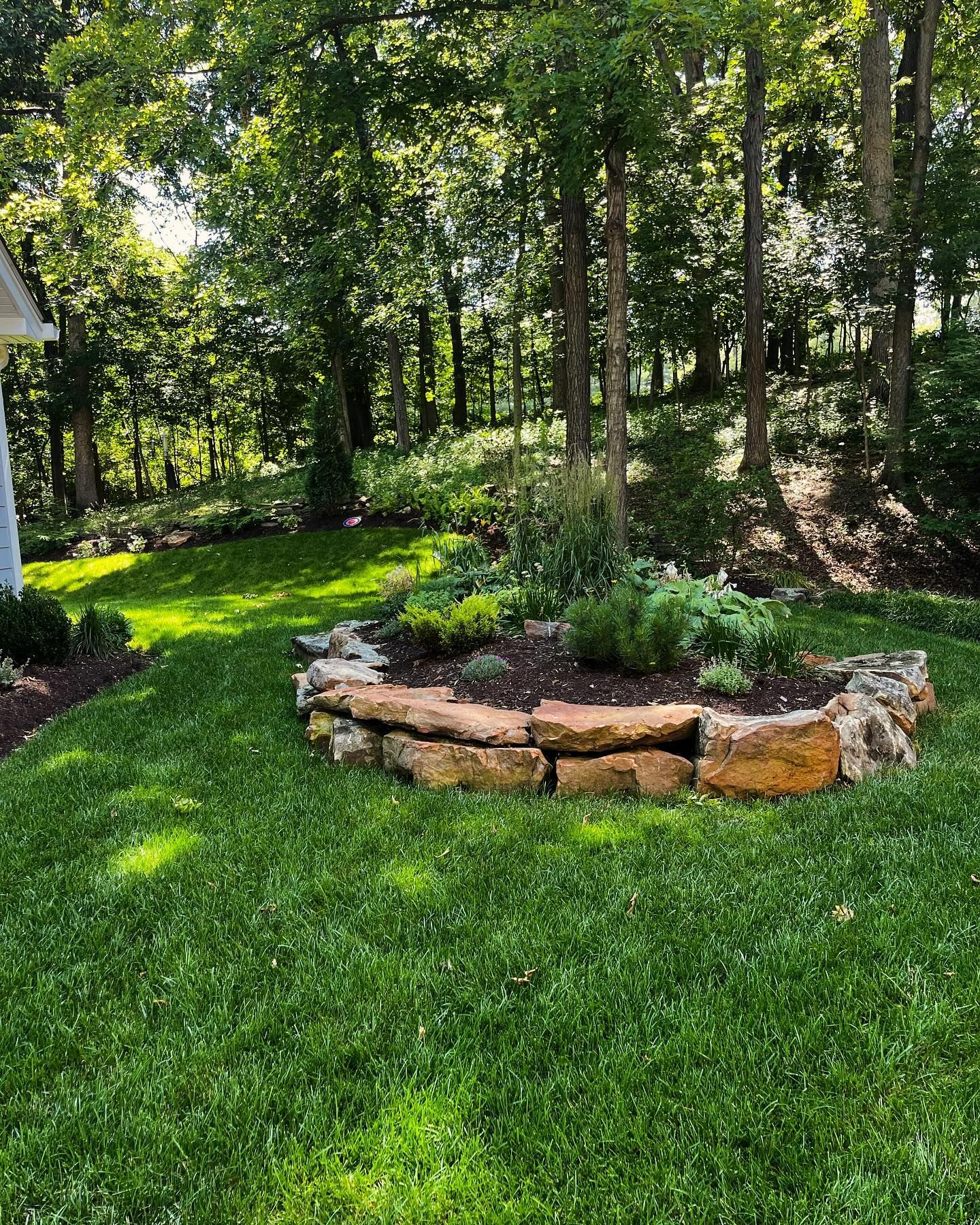 A circular garden bed made of stone is filled with plants, set in a grassy yard, with trees in the background.
