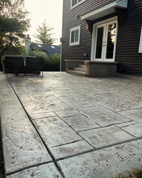 A stamped concrete patio in front of a dark-sided house with a wicker couch and double doors.