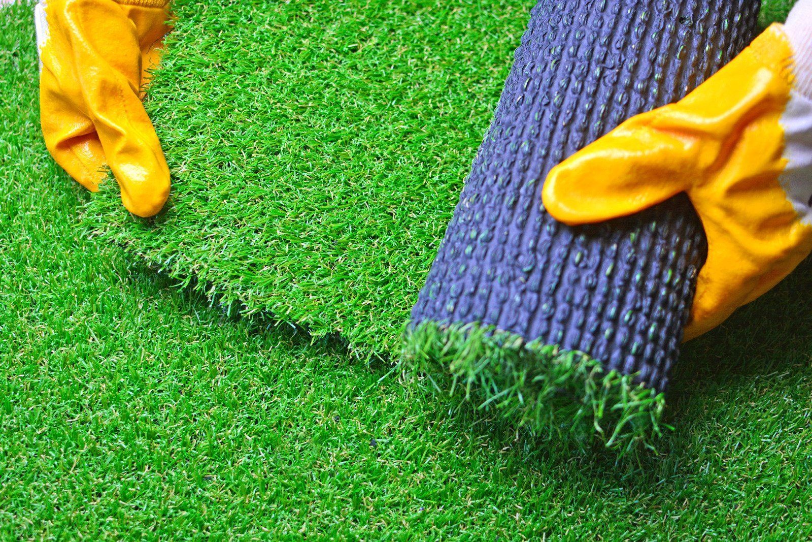 Person wearing yellow gloves unrolls a section of green artificial turf, revealing the black underside.