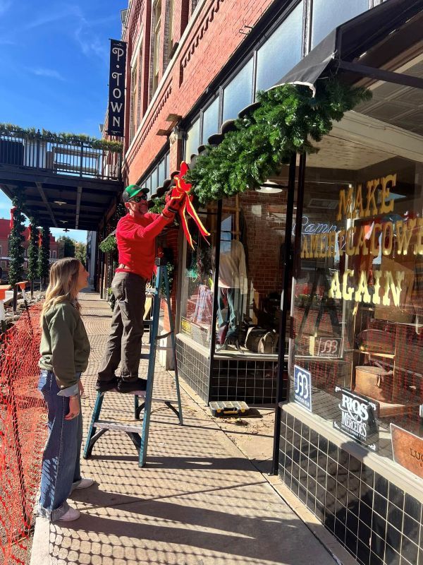 Man on ladder decorates a storefront with garland. A woman watches. Building in background.