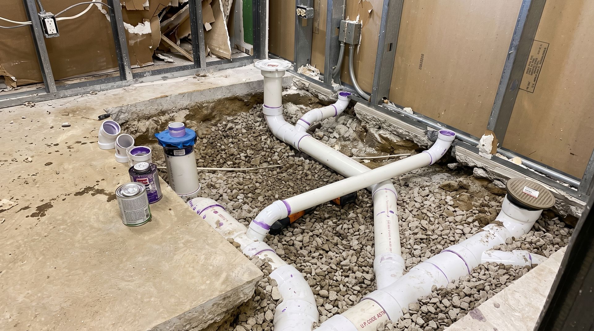 Plumbing pipes and fittings laid in a dug-up floor during bathroom renovation, with debris and tools nearby