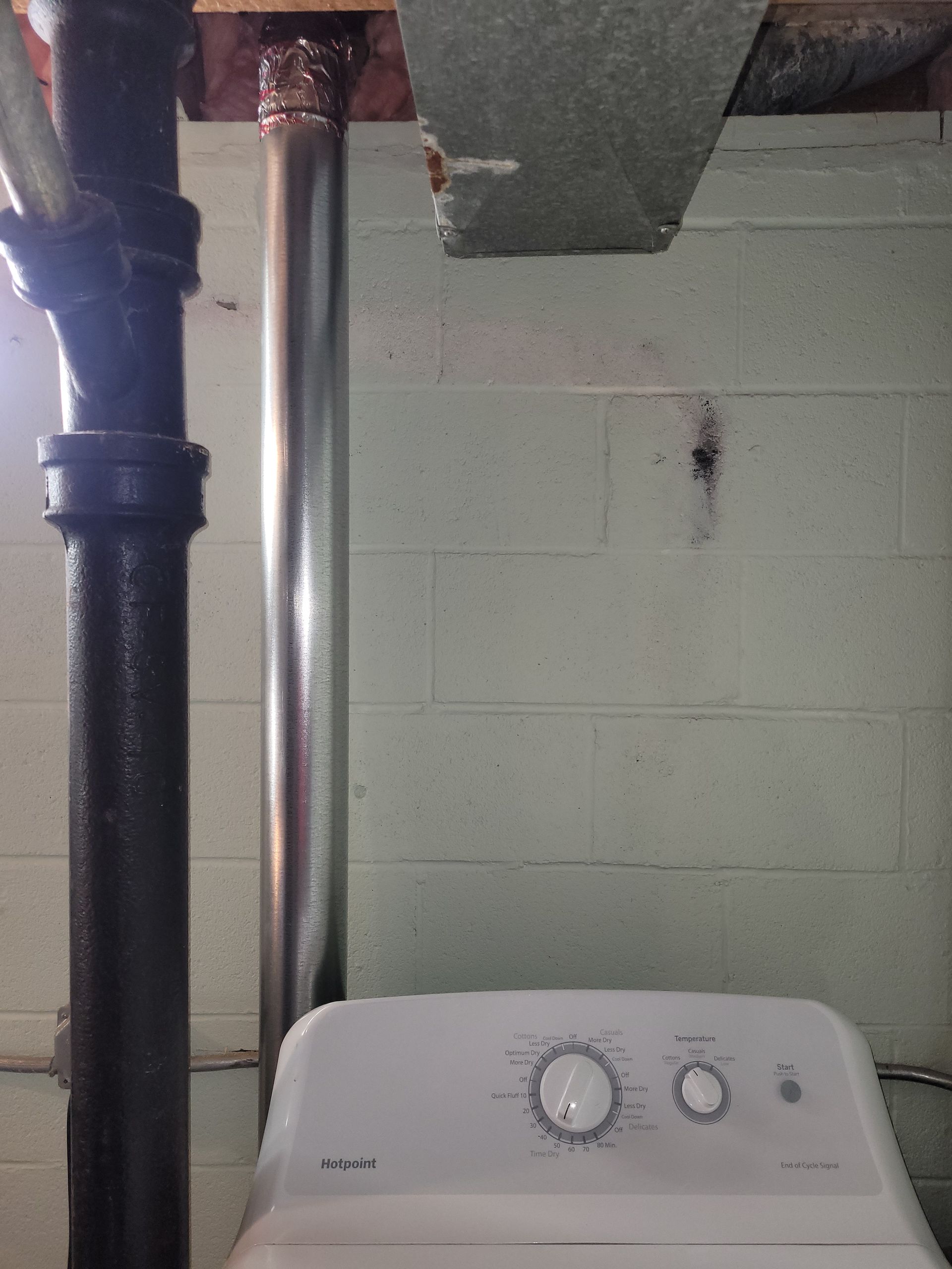 A dryer sits below a vent pipe and black water pipe against a cinderblock wall in a basement.