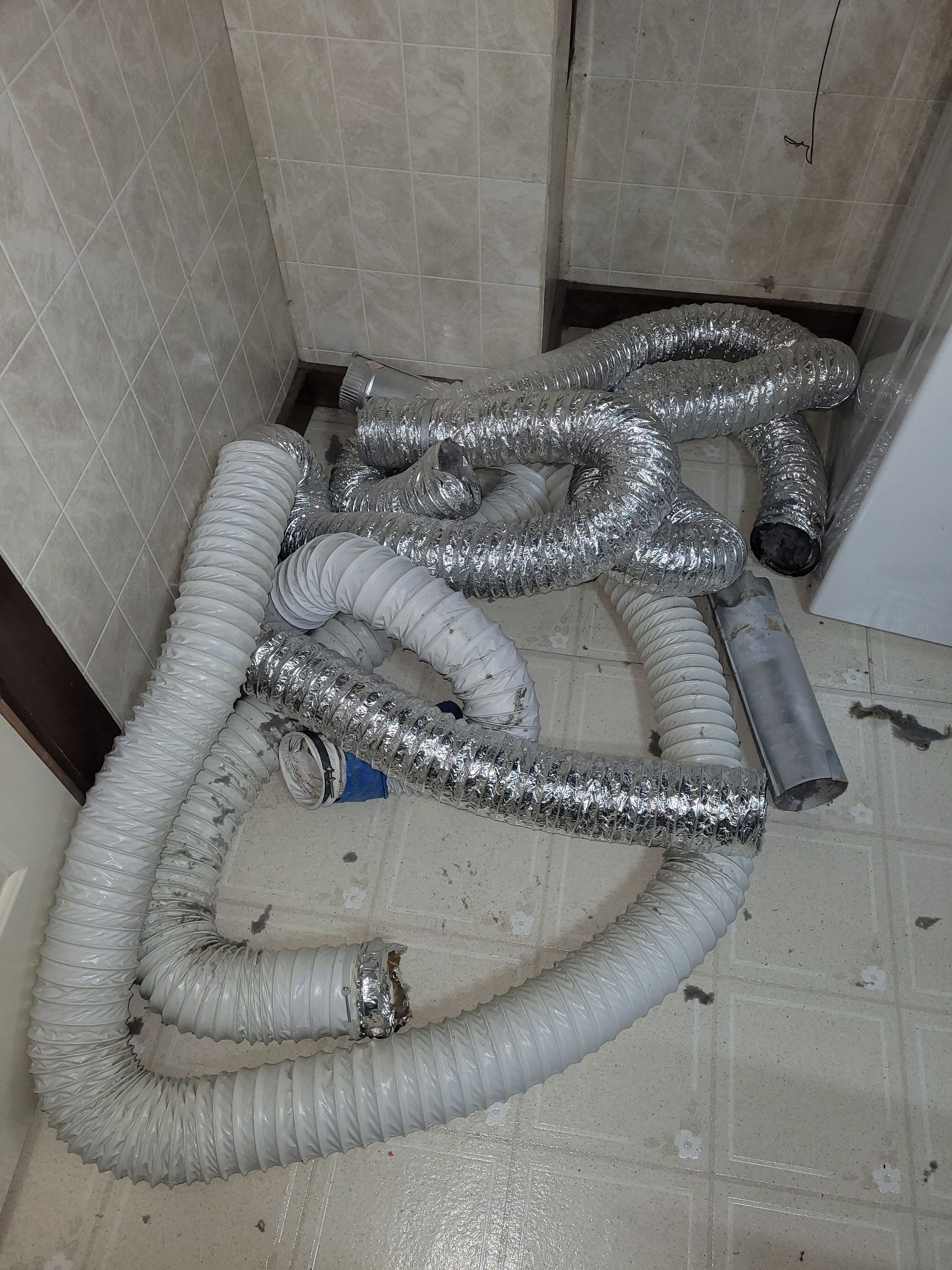Tangled, silver and white dryer vent hoses on a dirty floor in a corner.