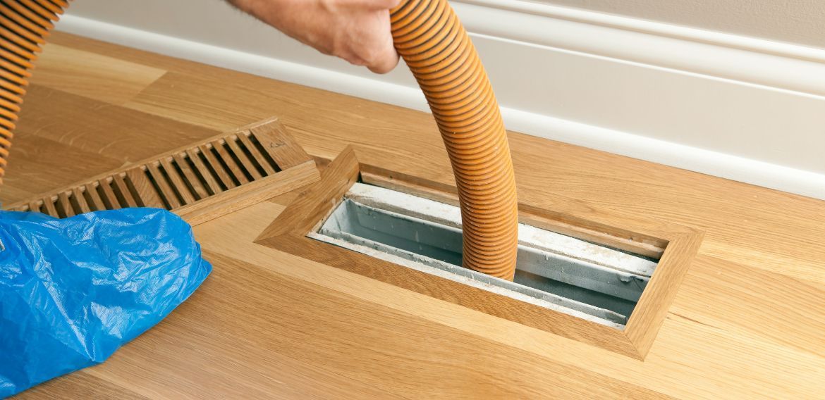 A person vacuums a floor vent with a corrugated hose.