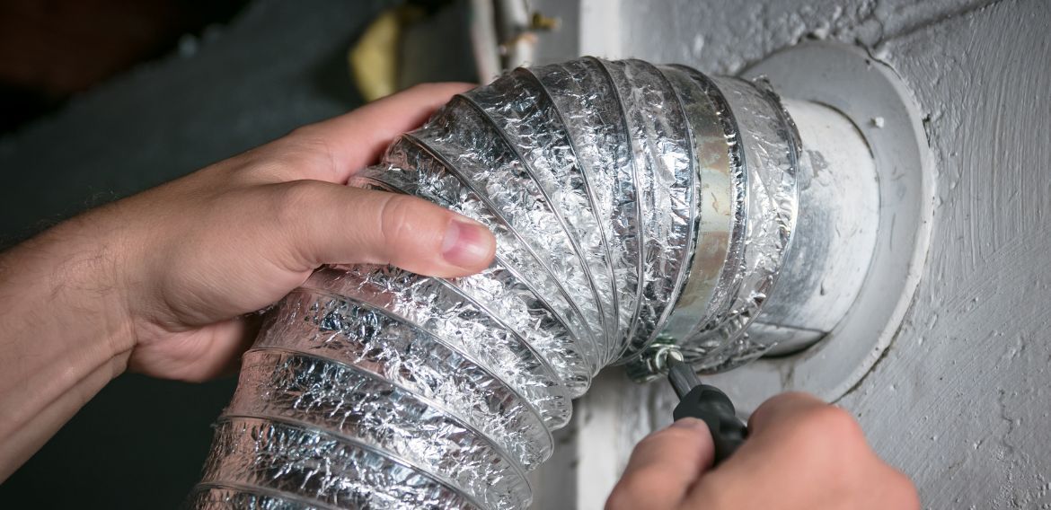 A person's hands attaching a metal dryer vent hose to a wall outlet.