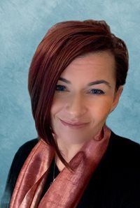 A person with short, reddish-brown hair smiles, wearing a black top and a shimmering copper-colored scarf.