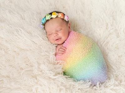 Sleeping newborn wrapped in a rainbow blanket with a flower headband on a fluffy white fur backdrop