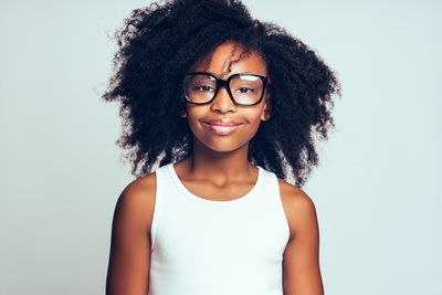 Smiling person with curly hair and black glasses wearing a white tank top against a light background