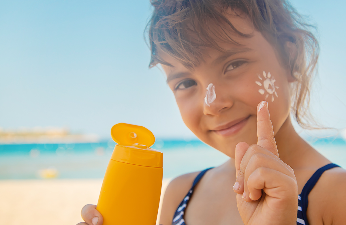 A person on a beach holds a yellow bottle of sunscreen while applying a sun-shaped dollop of cream to their cheek.