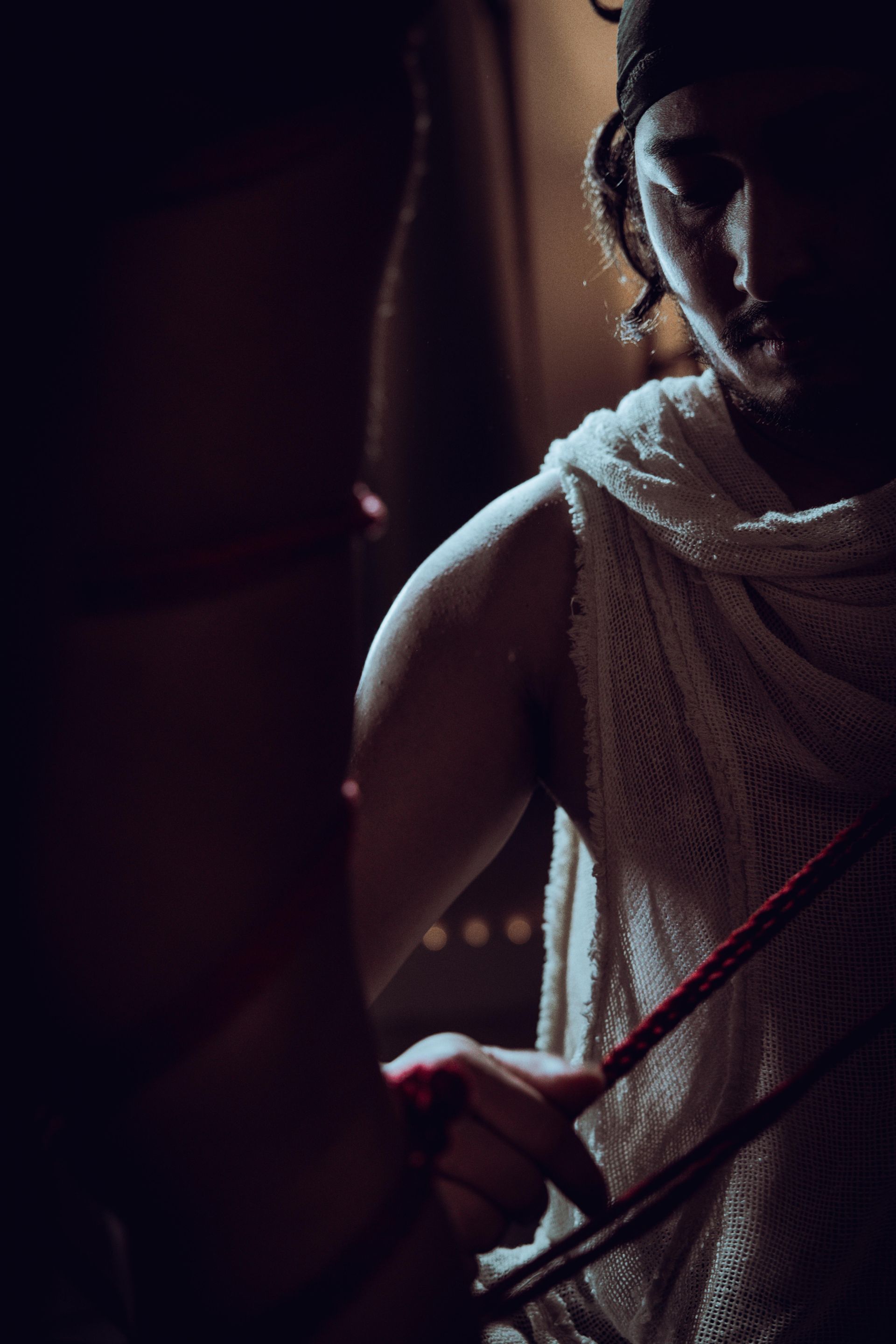 Man in white woven cloth holding red ropes, low-lit room, possibly preparing something.