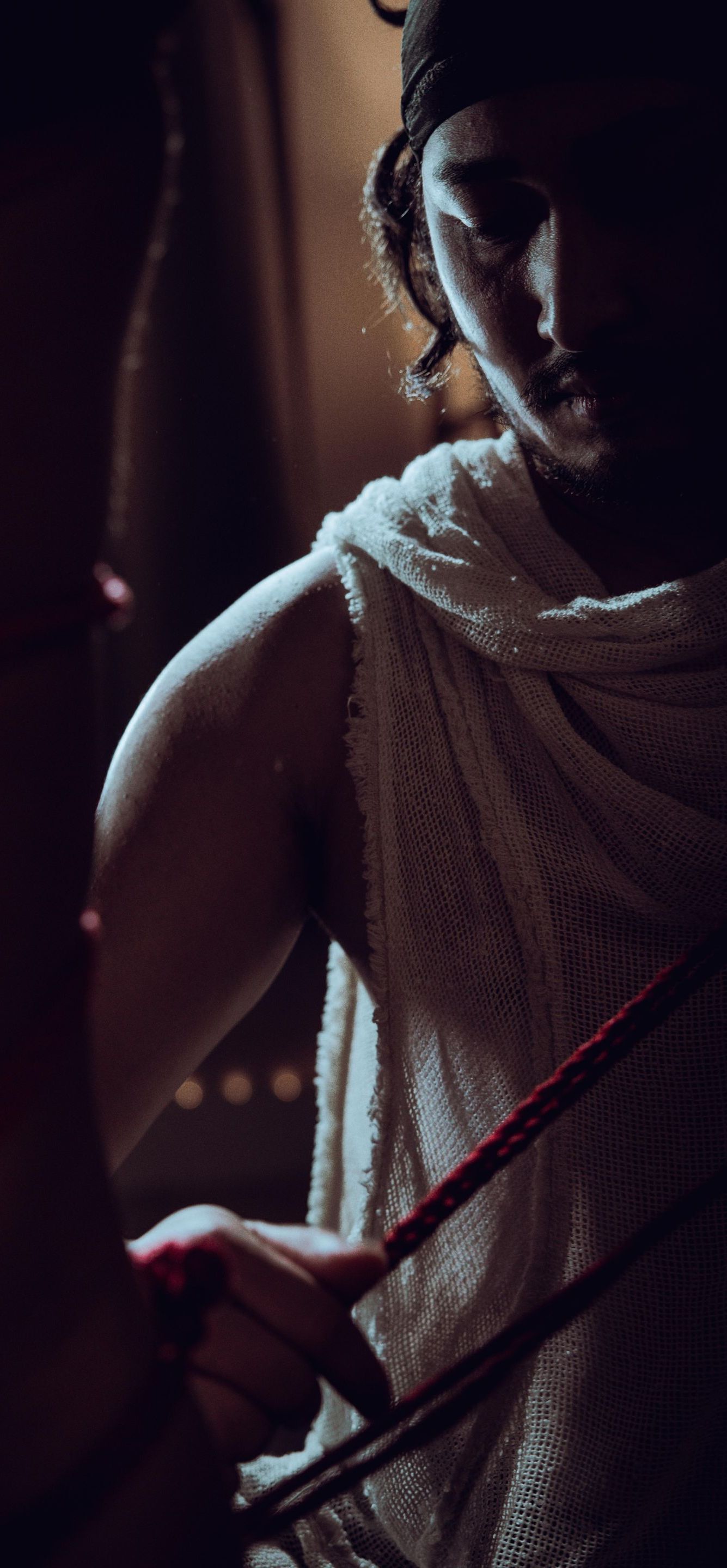 Person holding red ropes, wearing white draped cloth, arm flexed. Dark setting.