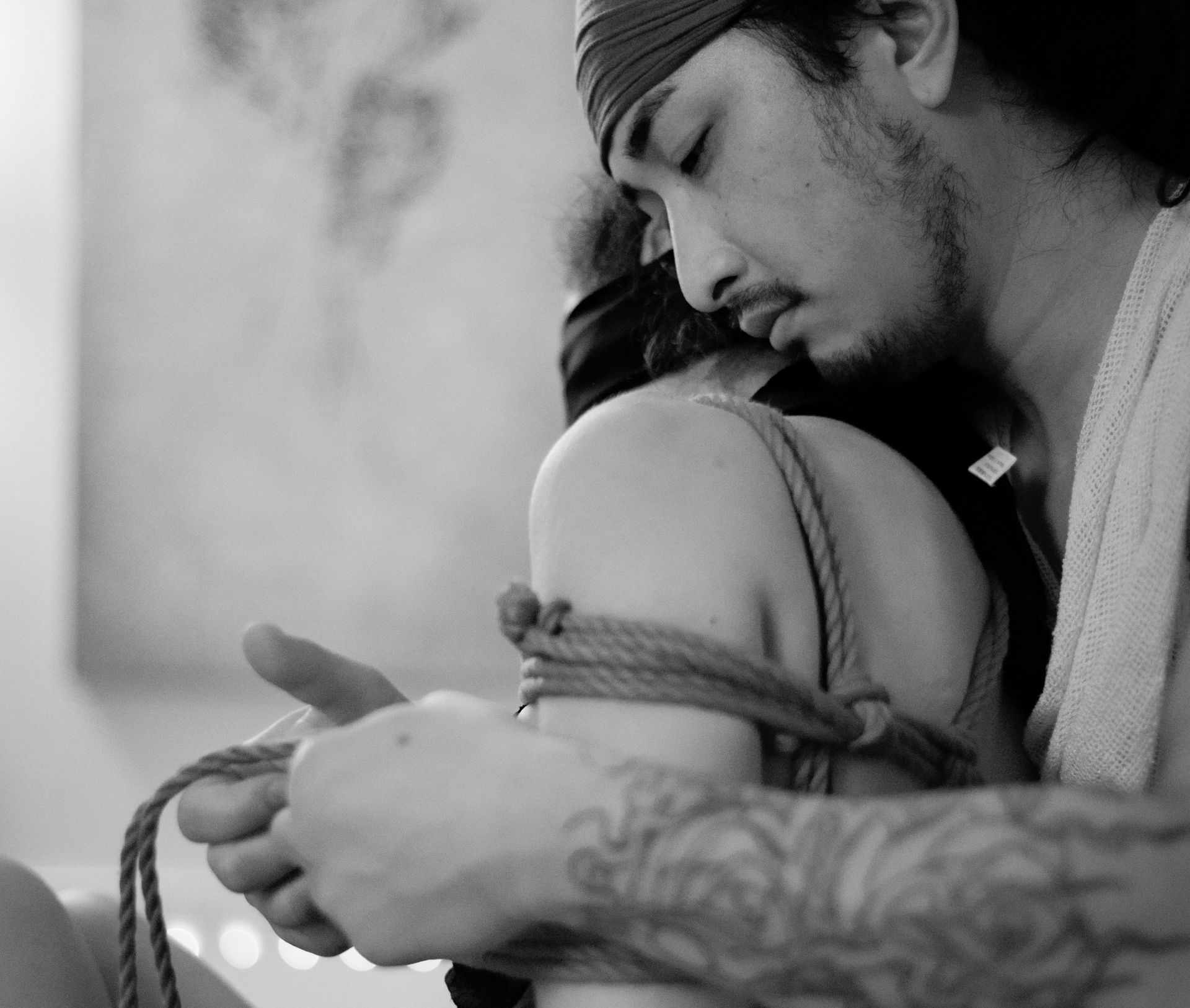 Man ties rope around a person's shoulder, face close, looking down. Black and white photo.