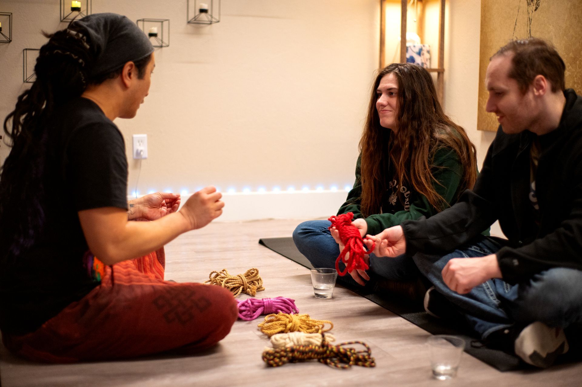 Three people seated on the floor, holding colorful string, smiling. Interior setting, well-lit.