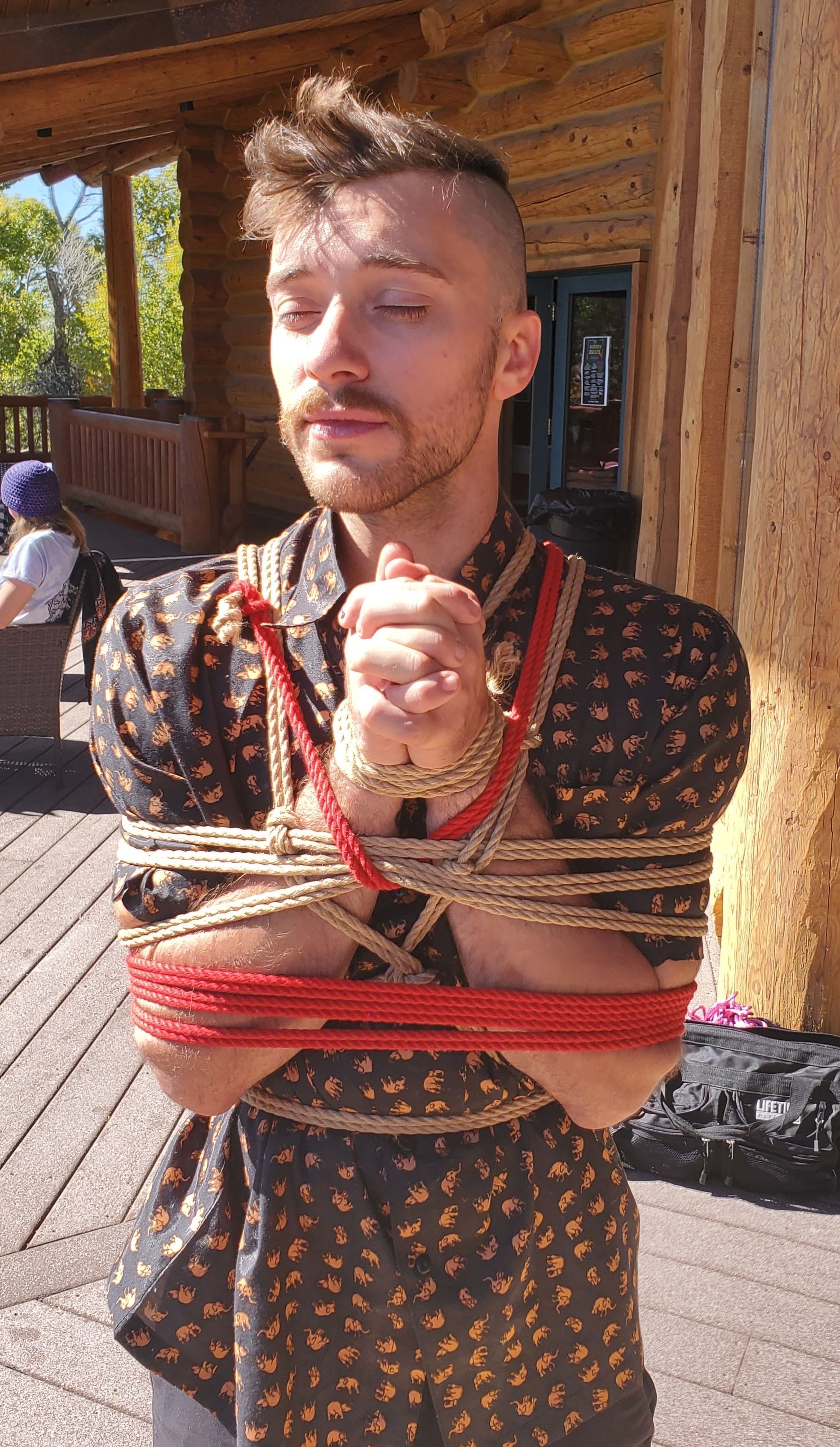 Man bound with rope, eyes closed, outdoors near a wooden structure.