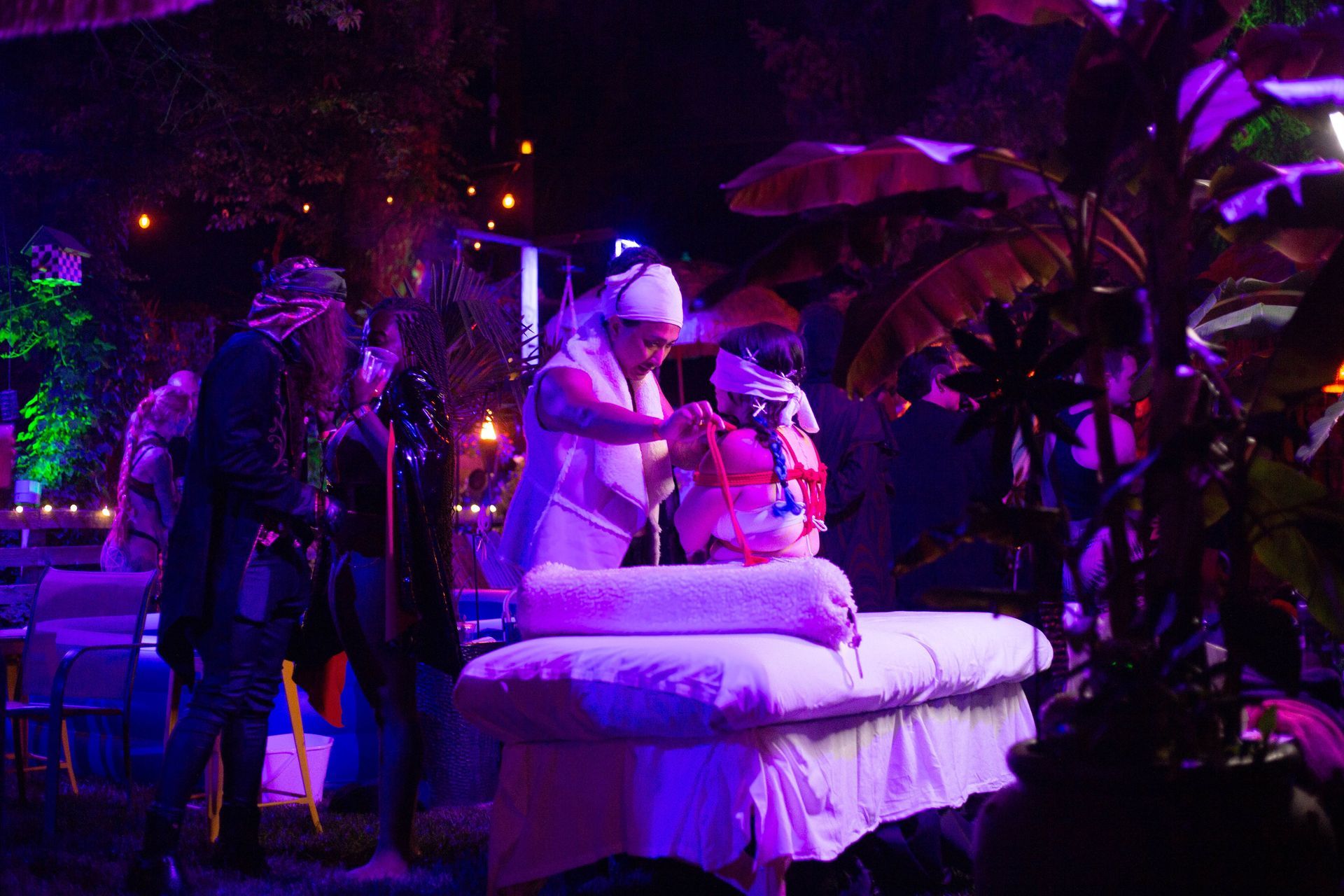 A person gives a massage to a person lying on a table outdoors at night, under colorful lights and plants.
