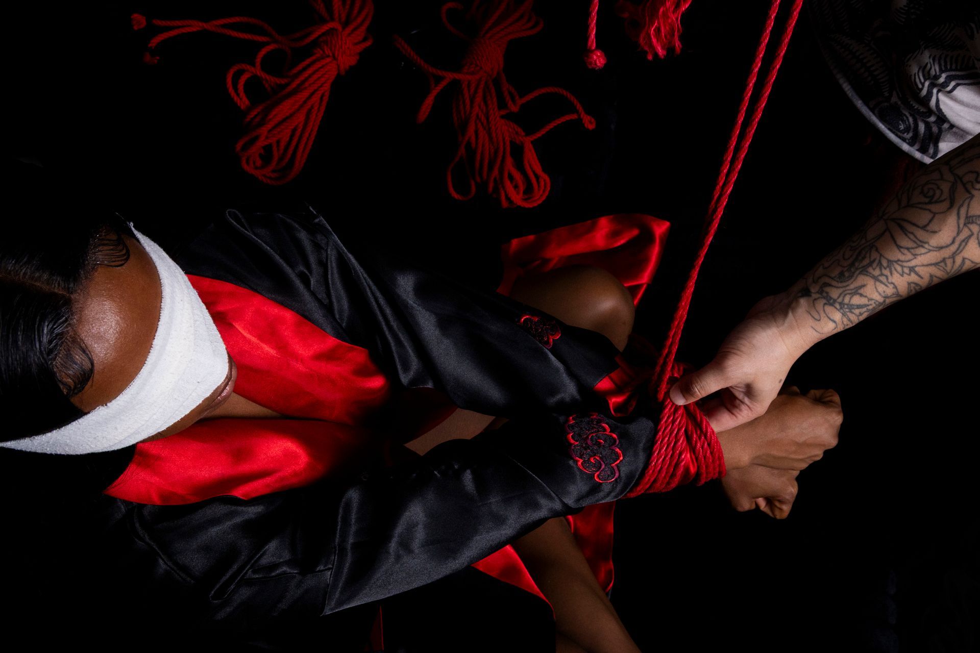 Person being tied up with red rope; black and red clothing. Hand with tattoo wraps rope. Dark background.