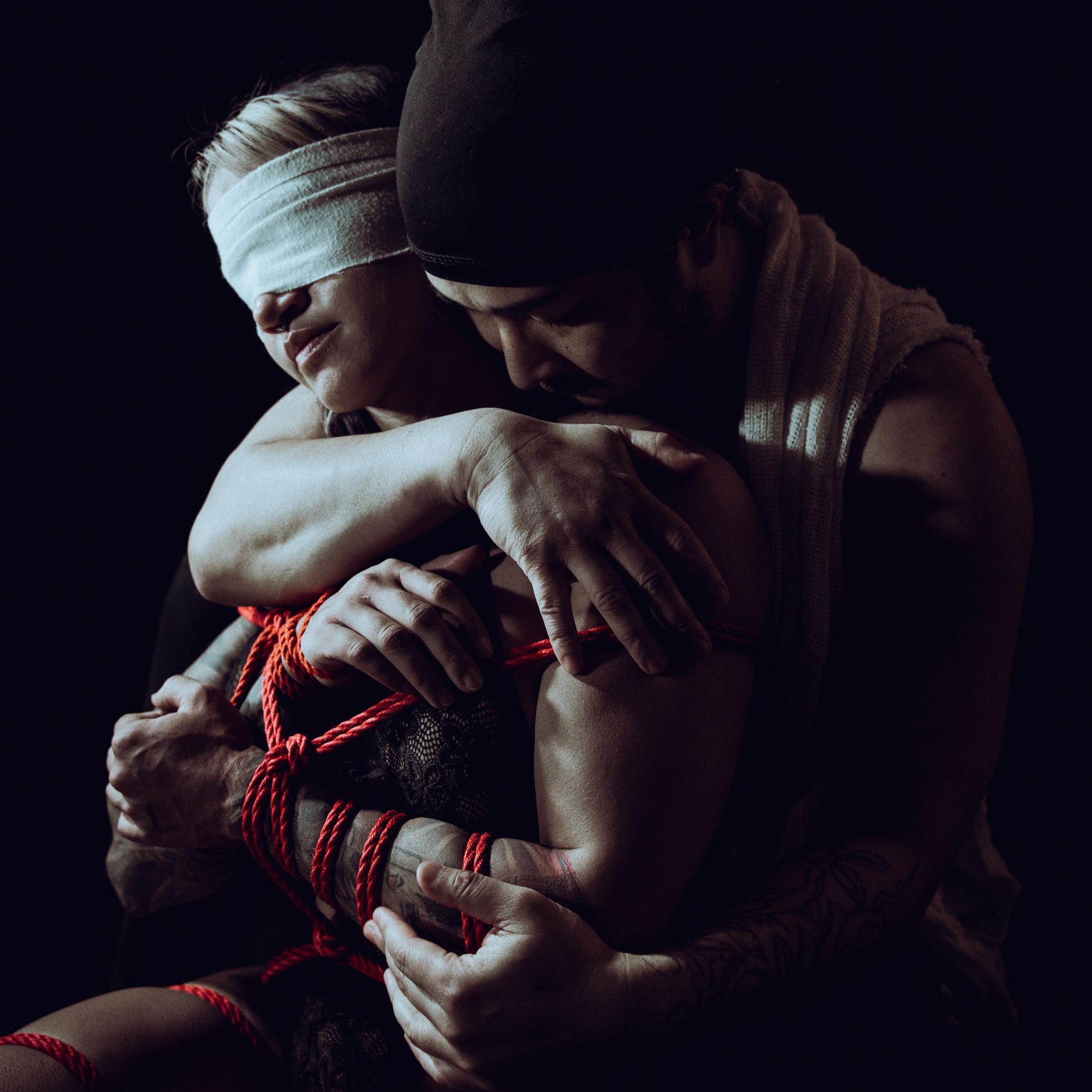 Person with blindfold tied with red rope, embraced by another person. Dark background.