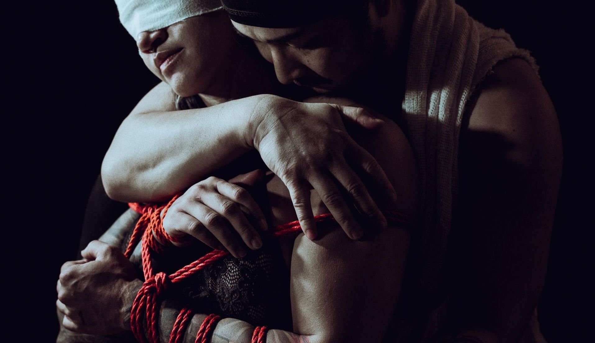 Person blindfolded, bound with red rope, embraced by another. Dark background.
