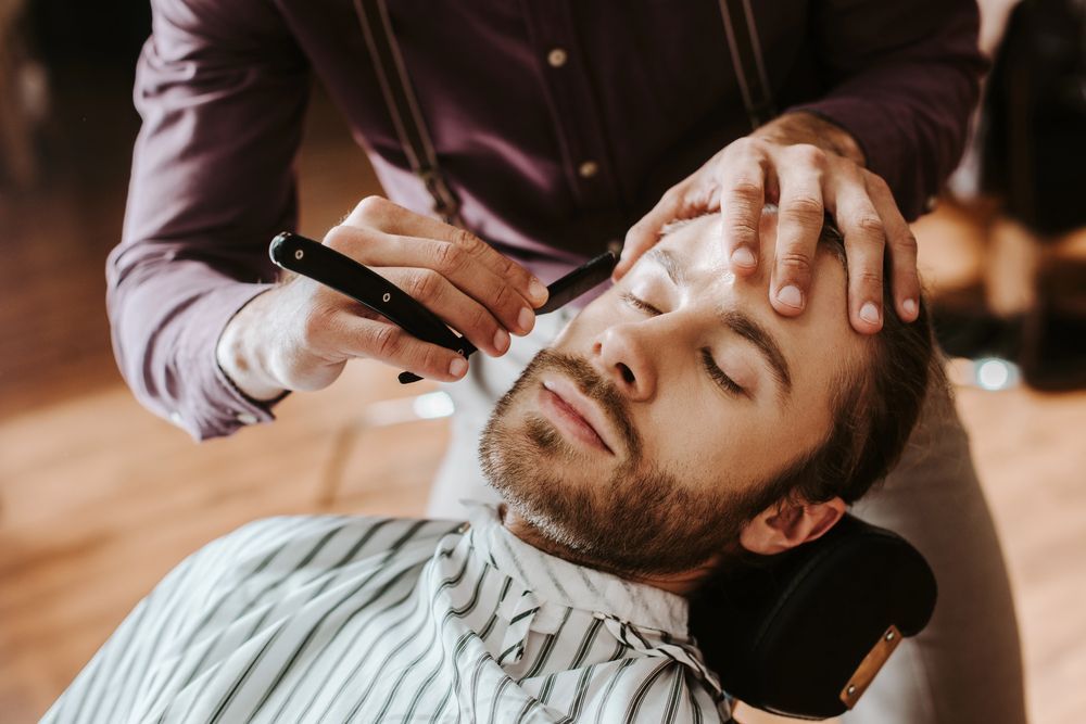Un barbero utiliza una navaja de afeitar para afeitar a un cliente relajado que está recostado en una silla de peluquería.