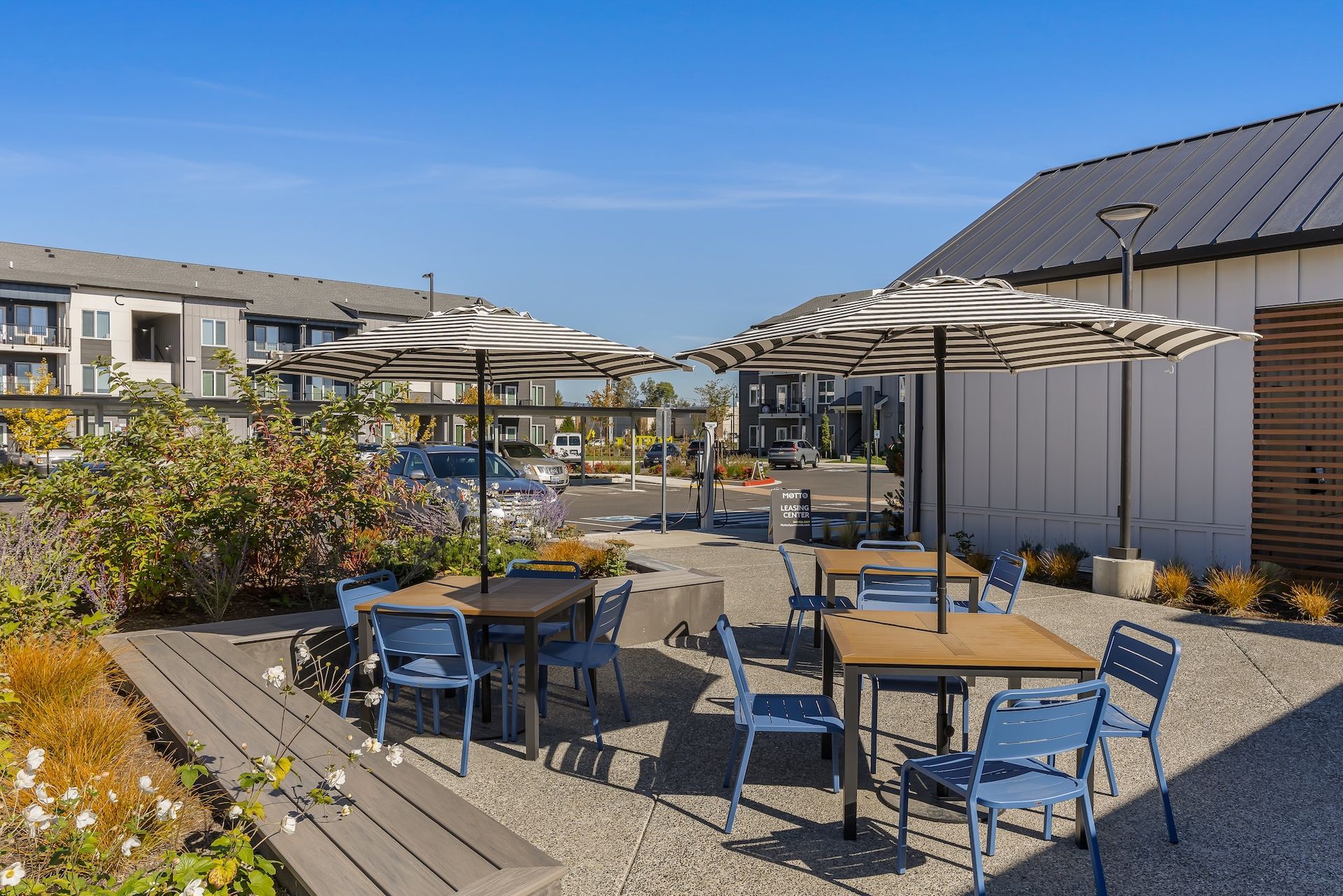 Outdoor patio with blue chairs, tables, and umbrellas beside modern buildings under a clear sky