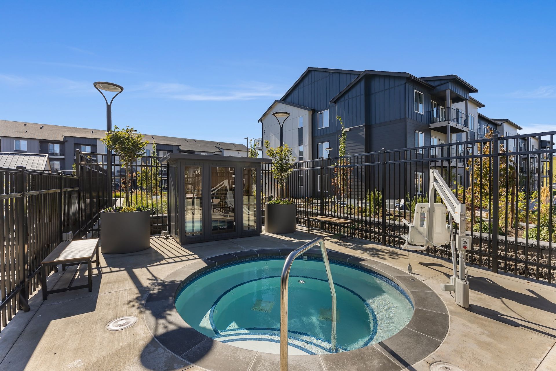 Courtyard hot tub beside a pool with black fencing and modern apartment buildings under a clear blue sky