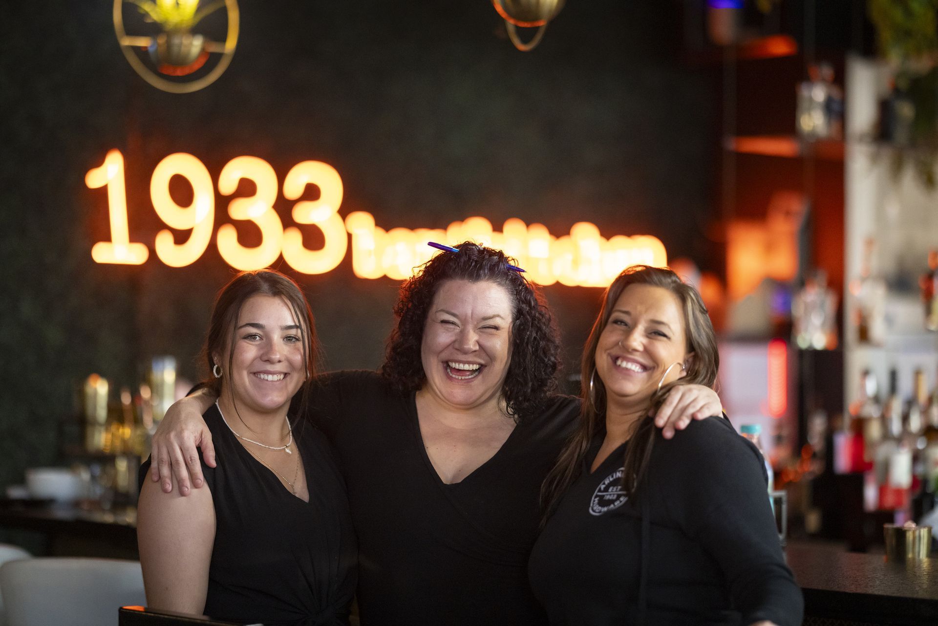 Three women are posing for a picture in front of a neon sign that says 1933.
