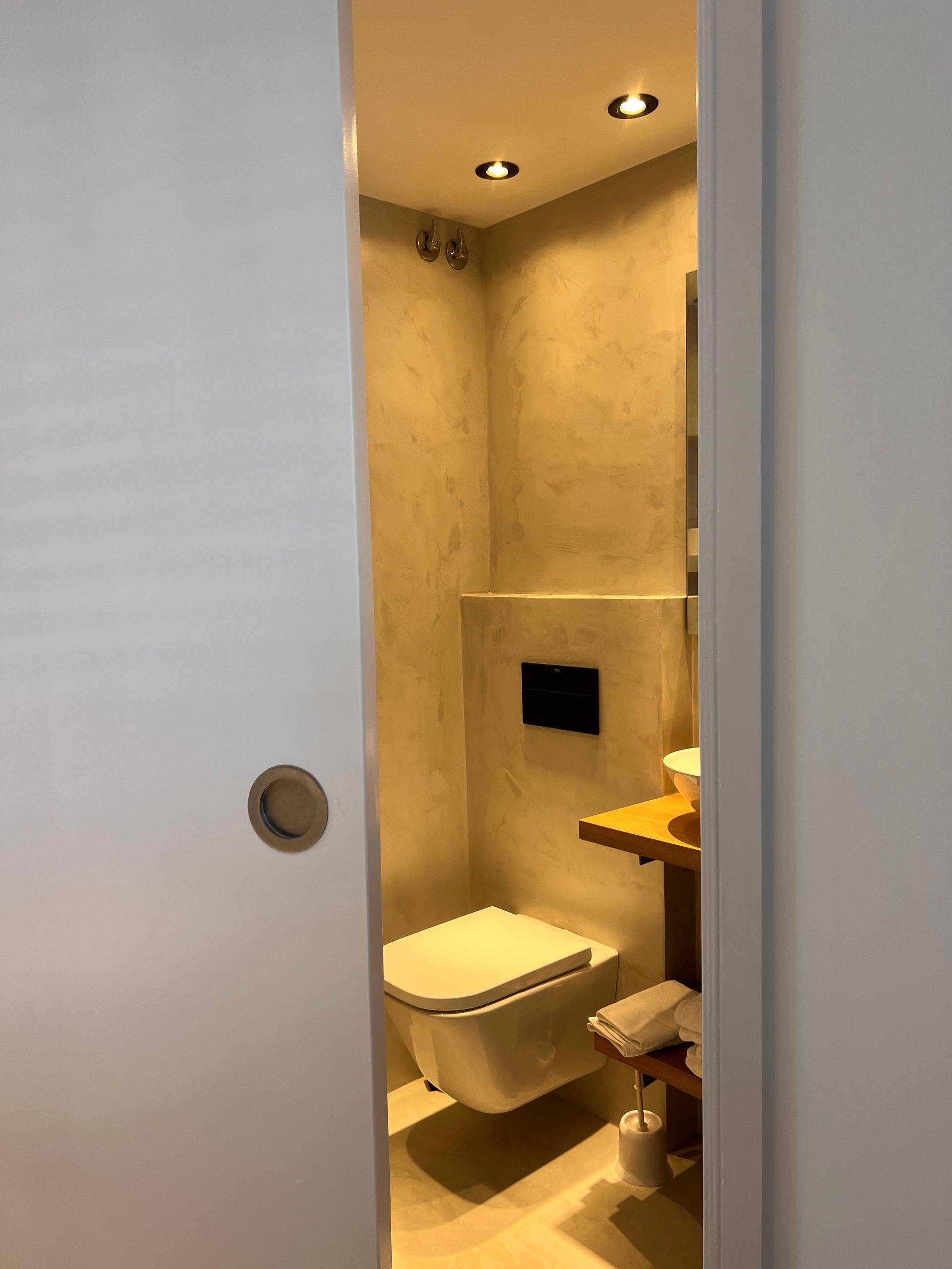 A narrow, modern bathroom with a wall-mounted toilet, wooden shelf, and recessed lighting.