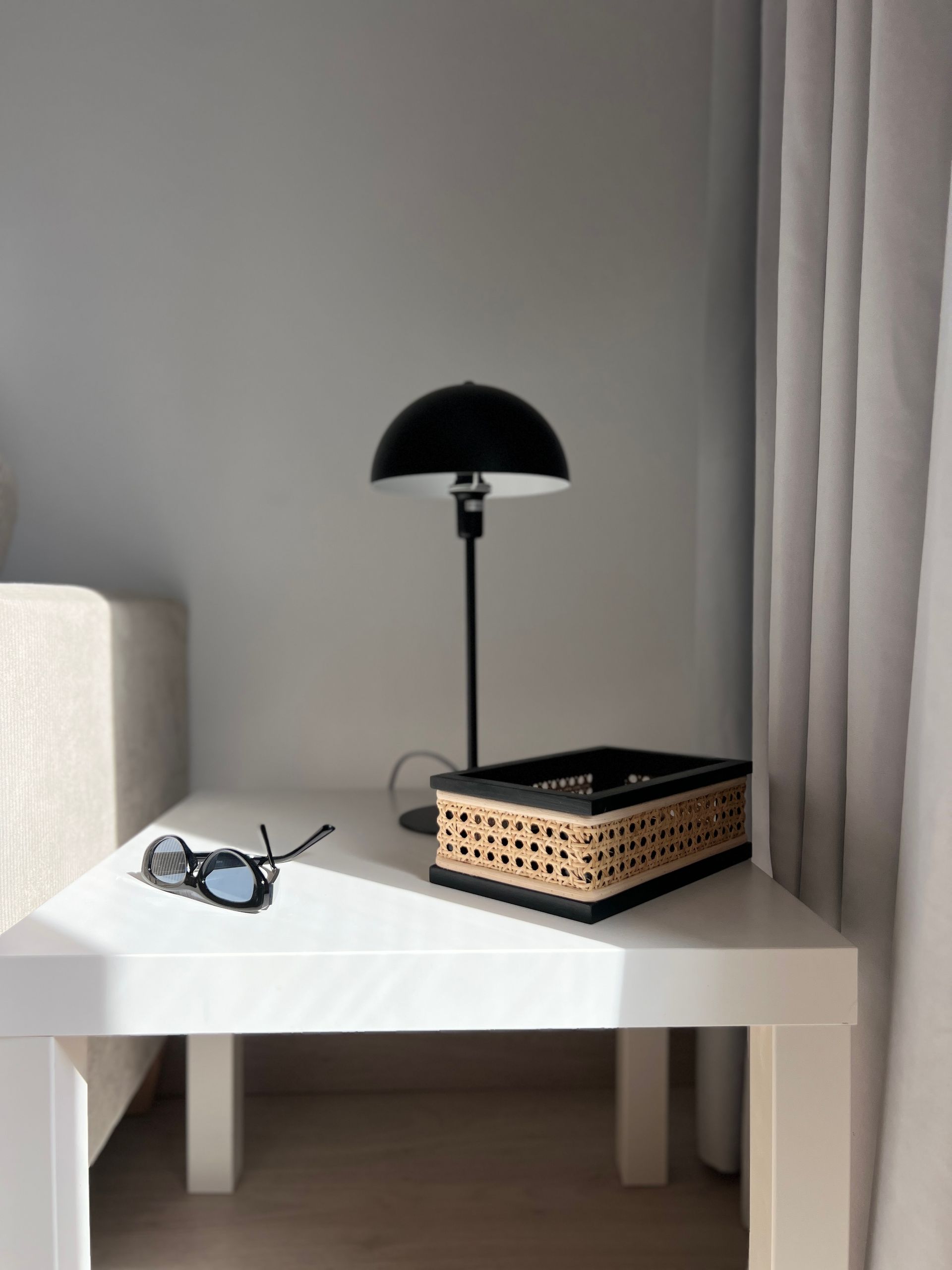 White table with black lamp, basket, and sunglasses. Next to a light-colored sofa and curtains.