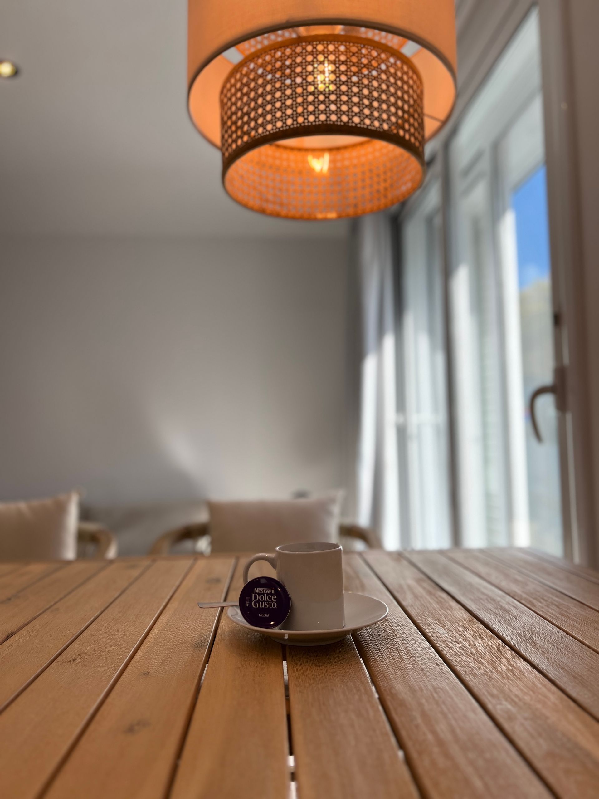 Cup of tea with a teabag tag on a wooden table, lit by a hanging lamp, French doors in the background.