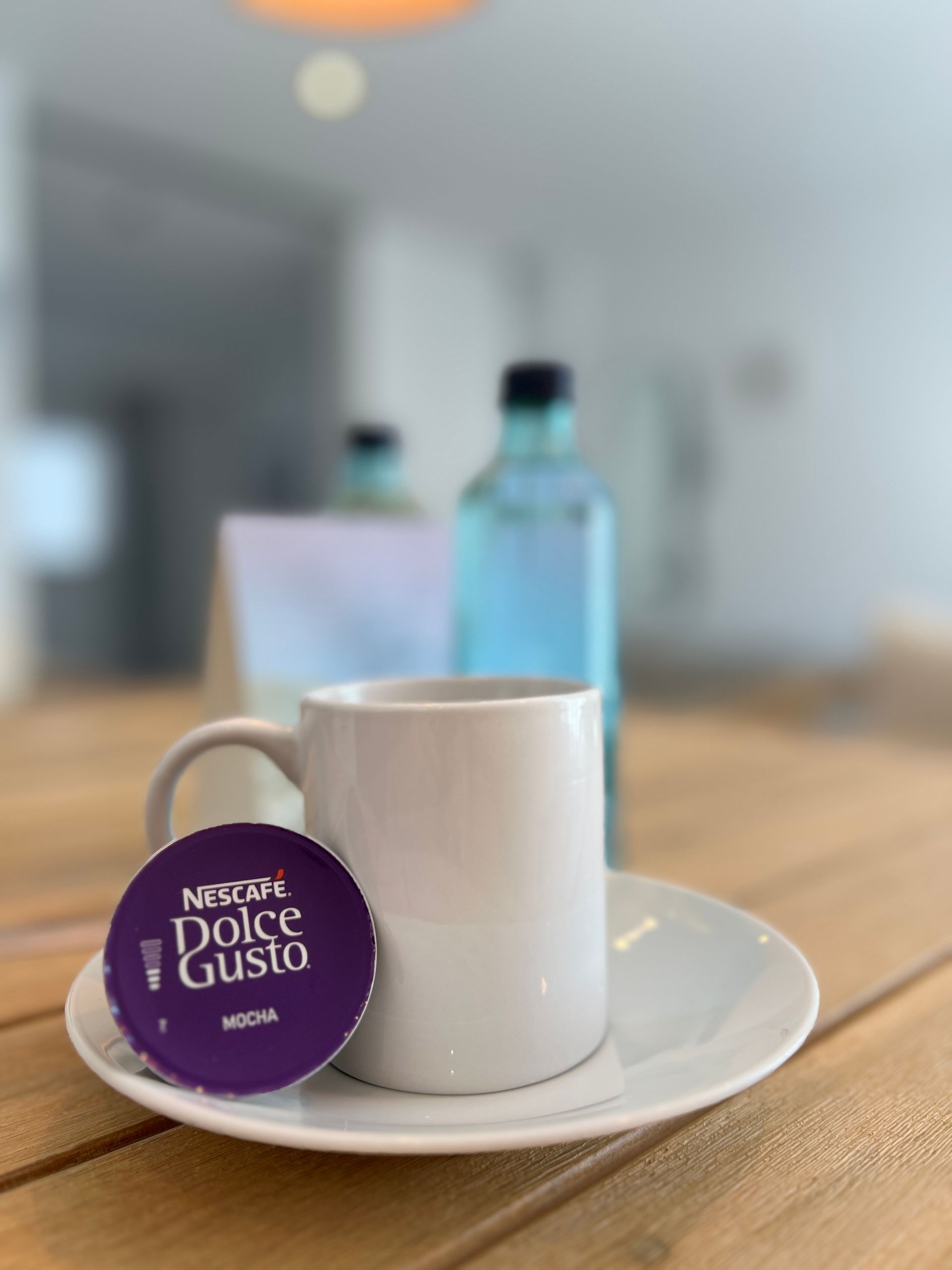 A white mug and Dolce Gusto pod on a saucer, with a water bottle and blurred background.