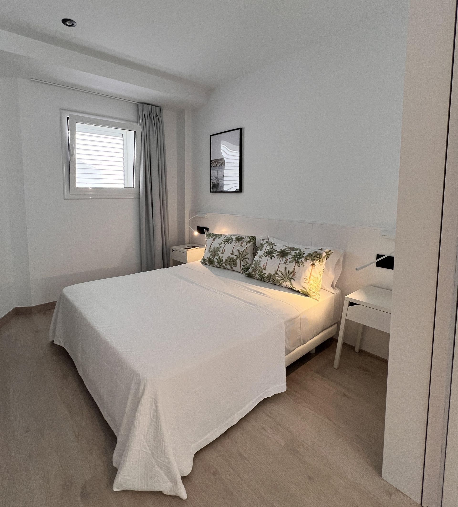Bedroom with a bed, white walls, wood floors, small window with curtains, and artwork.