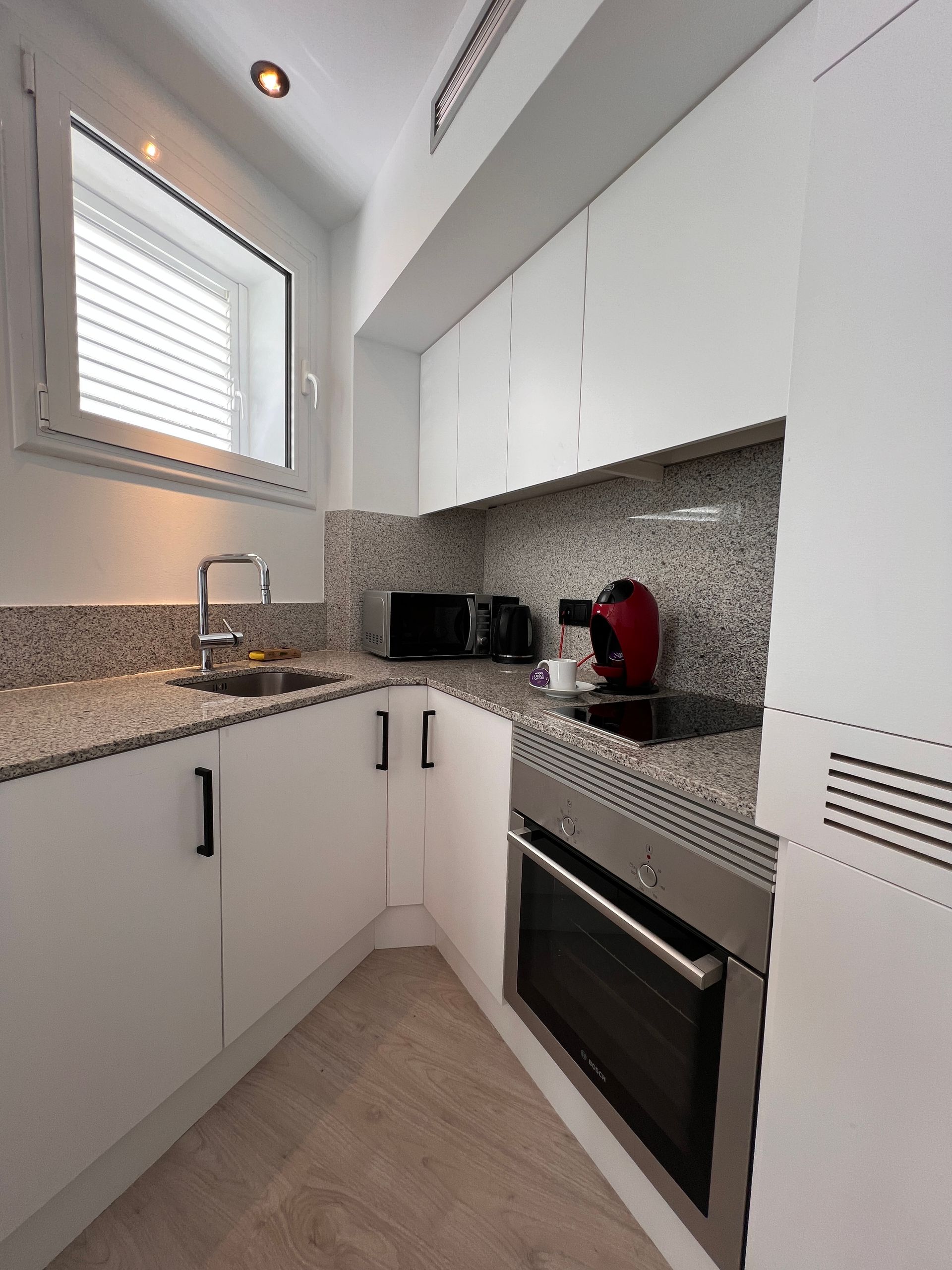 Small, white kitchen with granite countertops and stainless steel appliances. Window with blinds on the left.