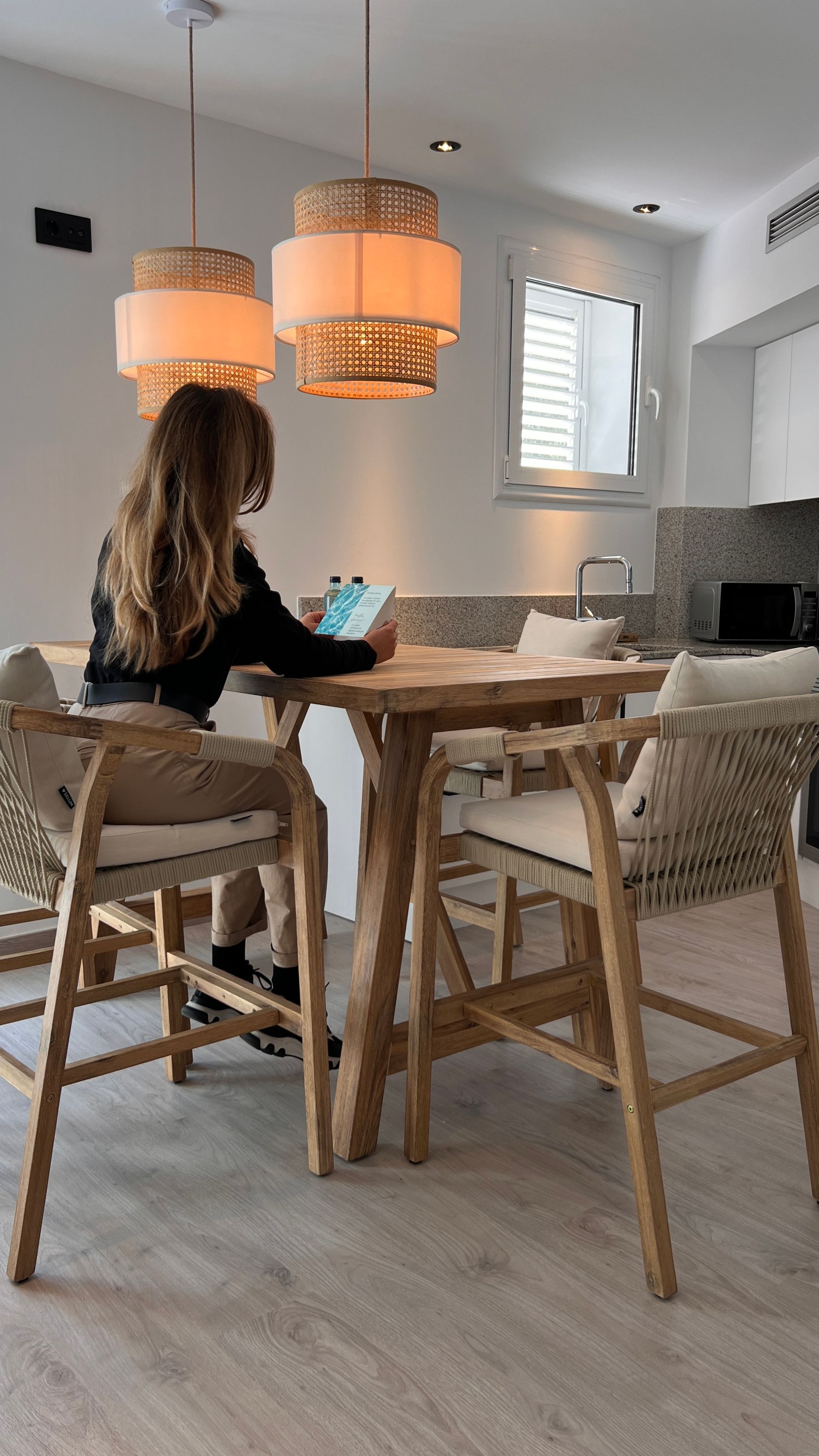 A person sits at a wooden table, using a tablet, under two pendant lights.