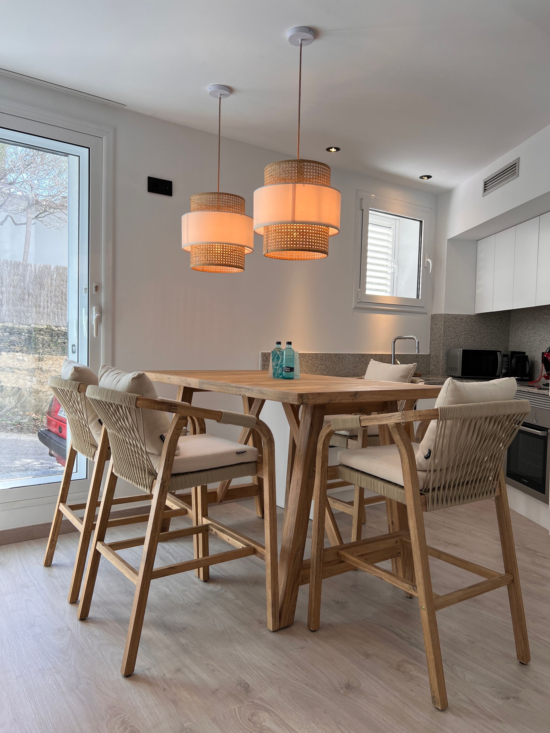 Round wooden table with four woven-backed bar stools beneath two pendant lights in a kitchen setting.