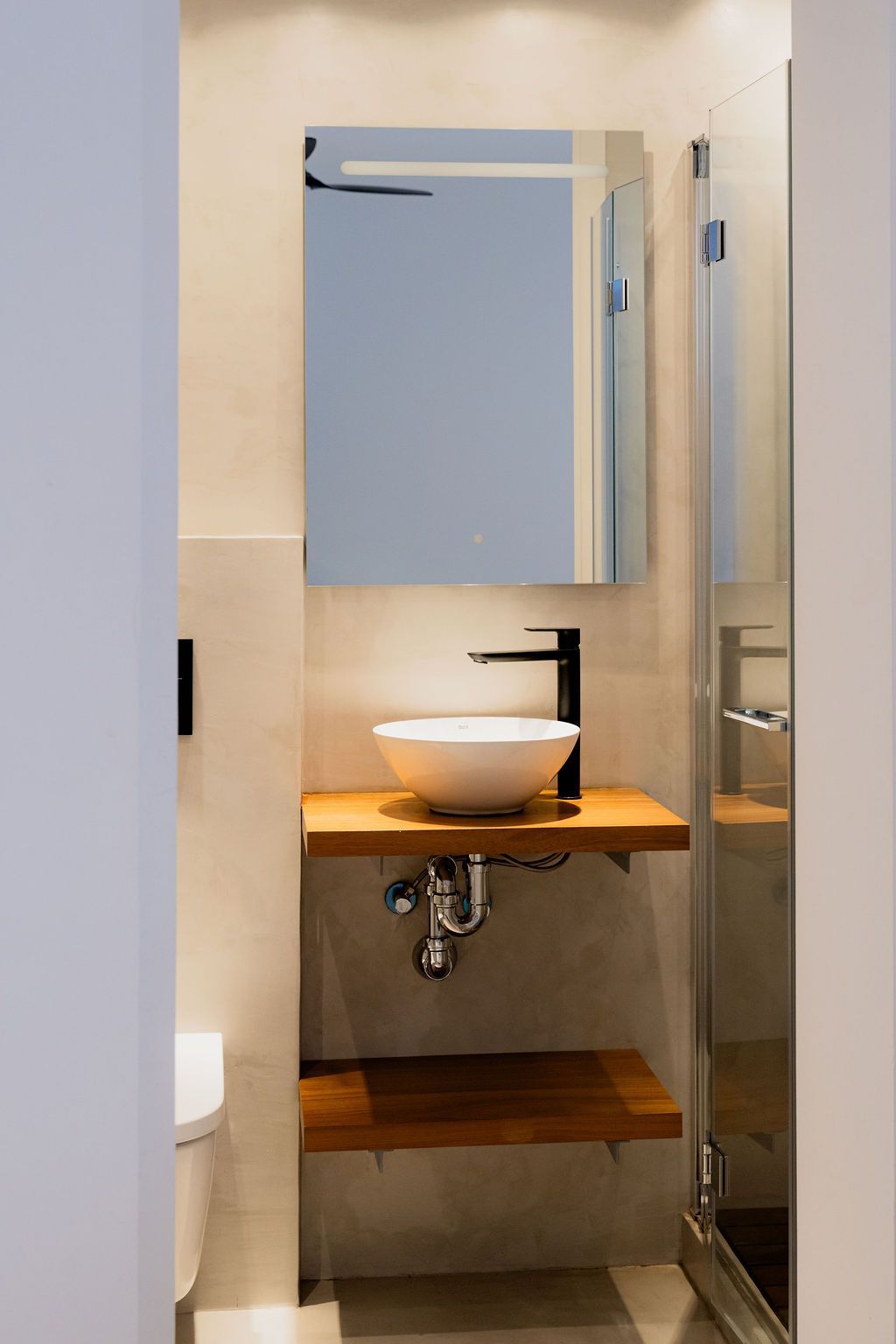 Small bathroom with white sink on a wooden shelf, black faucet, mirror, shower door.