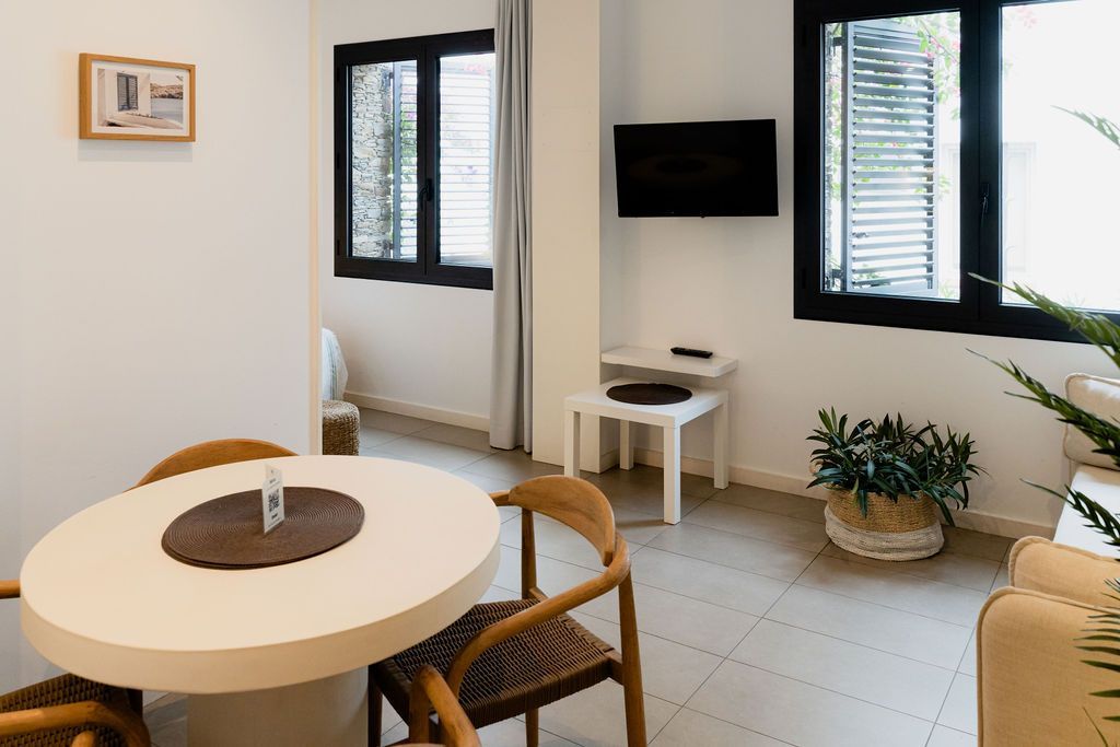 Bright living space with round table and chairs. Two black-framed windows, TV, white walls, and a plant.