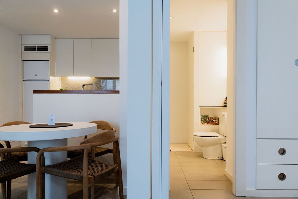 Kitchen and dining area with a view of a bathroom through an open doorway. White cabinets, wood chairs, and a round table.
