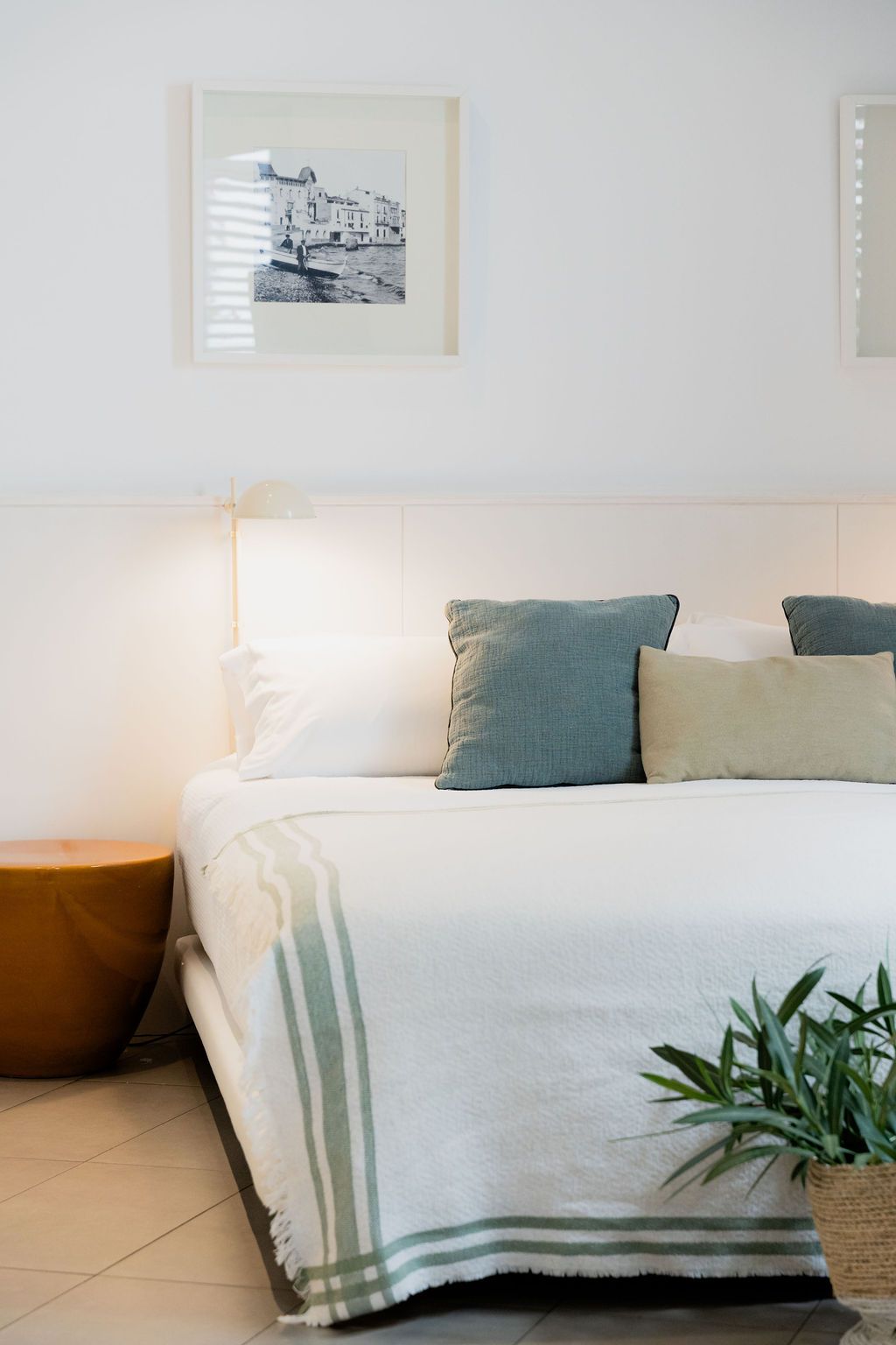 Bedroom with white walls, bed with pillows, blanket, painting, and a small plant.