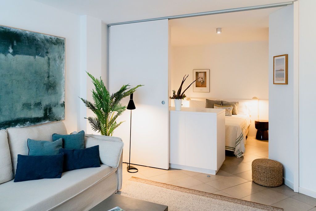 Modern living room with open door to a bedroom, featuring a sofa with blue pillows, a tall plant, and a bed.