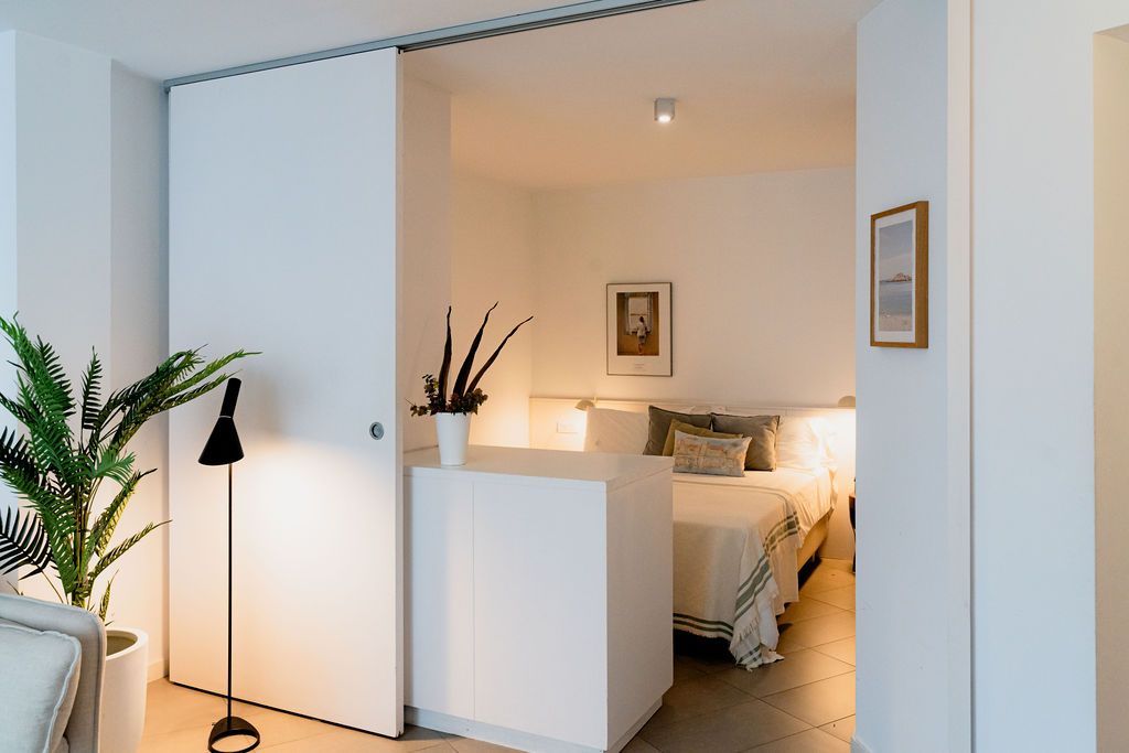 White bedroom with sliding door open to the bed. Plants and art on the walls.