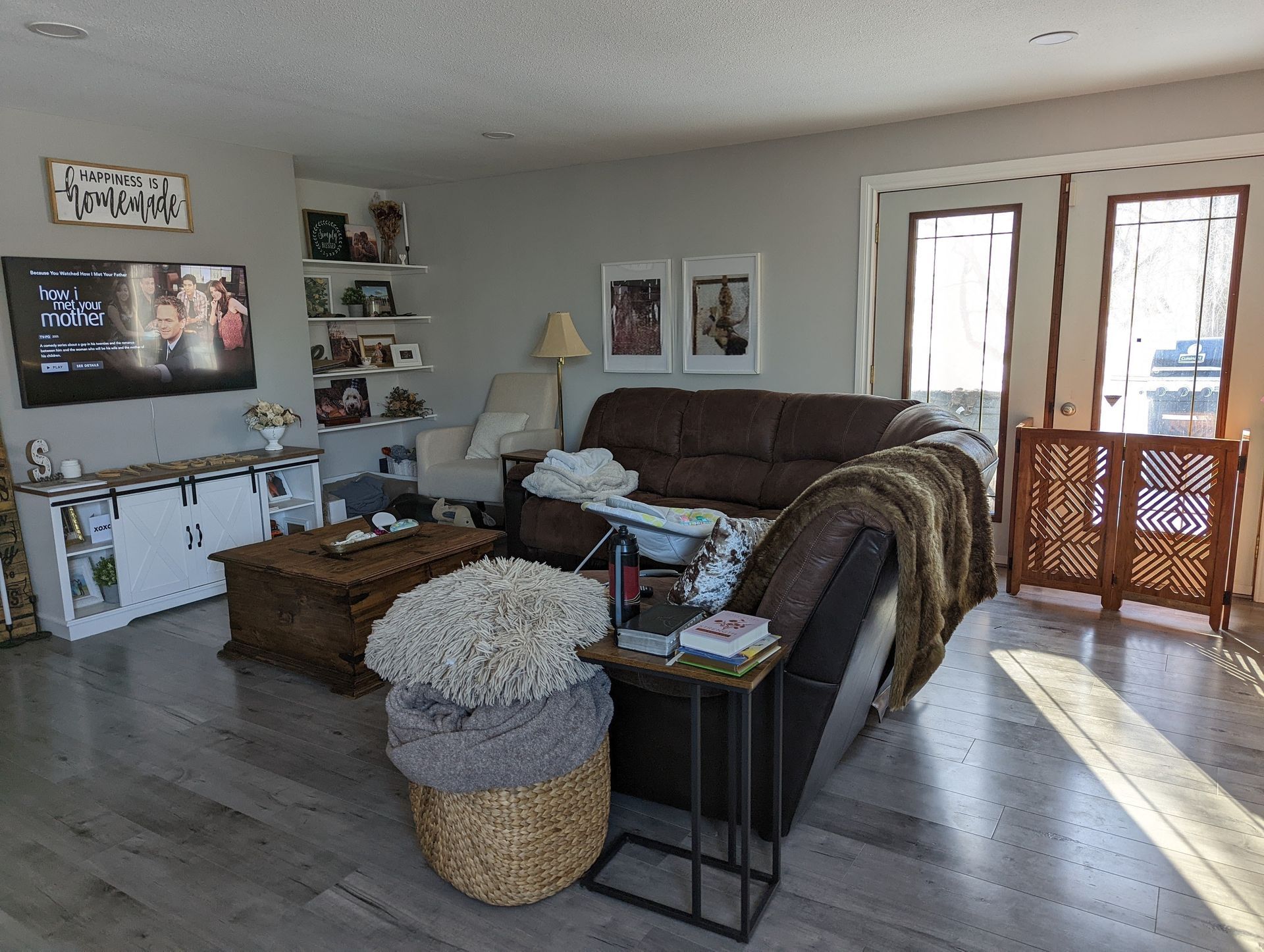 Living room with couch, TV, shelves, and wood floors. Sunlight streams through window.